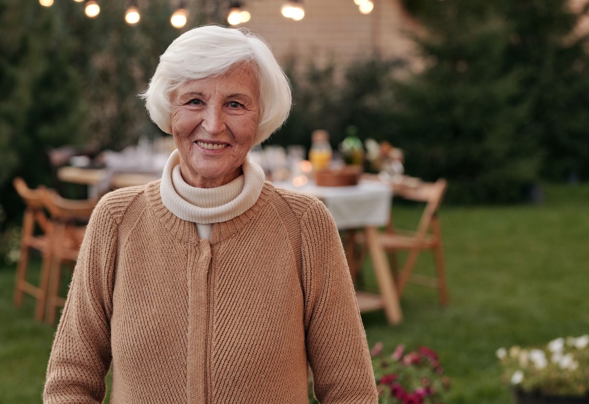 Lächelnde Seniorin steht im Garten, im Hintergrund ein gedeckter Tisch