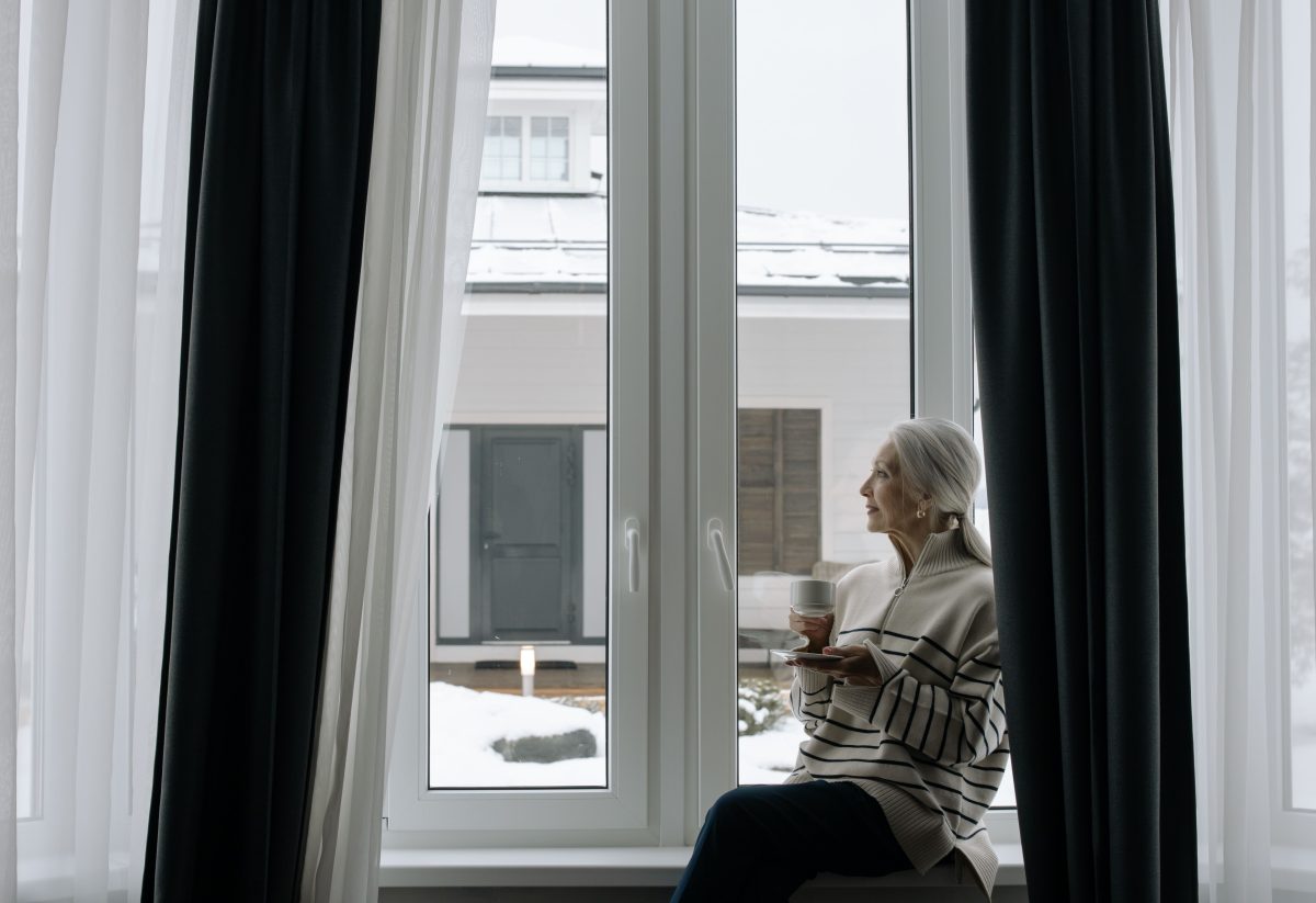 Seniorin sitzt mit einer Tasse am Fenster ihrer Wohnung und schaut nach draußen