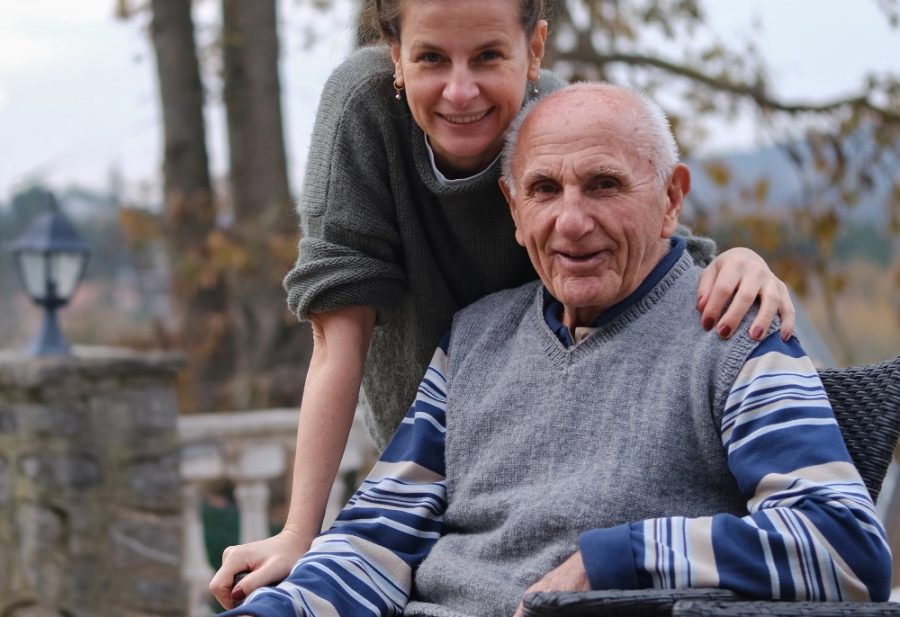Betreuungskraft steht hinter einem Senior, der auf einer Bank im Garten sitzt