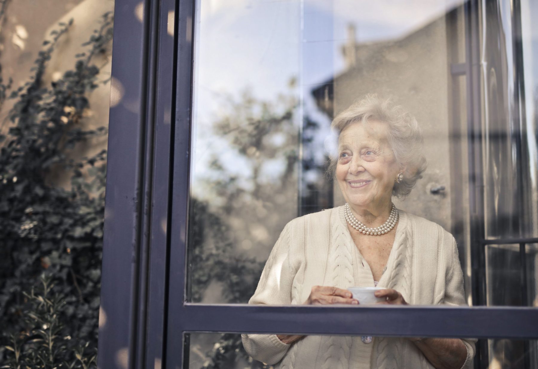 Seniorin steht mit einer Tasse am Fenster ihrer Wohnung und blickt in den Garten